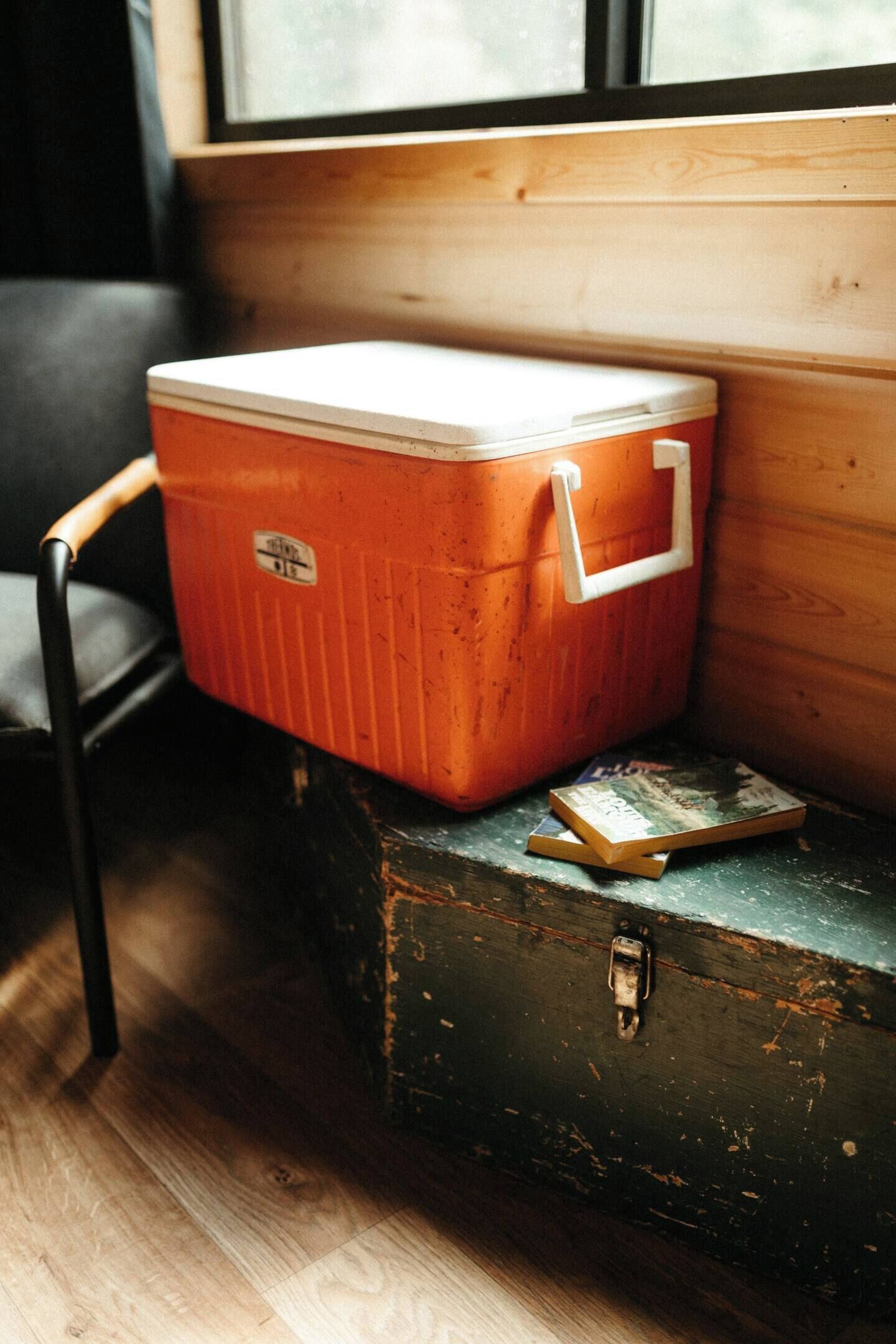 A vintage orange cooler with a white handle resting on a wooden bench next to a black suitcase.