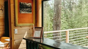 Interno di una baita in legno con angolo bar, sgabelli verdi, un cartello di benvenuto incorniciato e una finestra vista foresta.