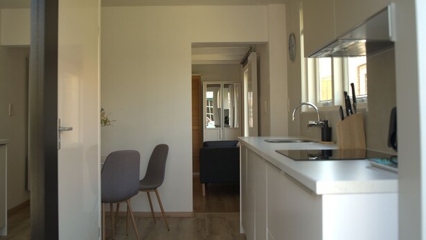 A narrow hallway with a kitchen area on the right, featuring a sink, countertop, and wooden chairs.