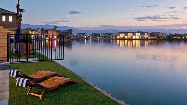 A waterfront property with a pool, lounge chairs, and a view of the mountains. - Luxe Lakefront Dream Villa: Heated Saltwater Pool (Indio)