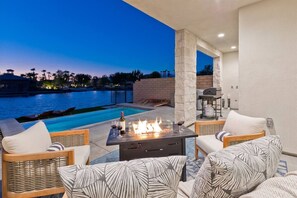 An outdoor patio with a fire feature, seating, and a view of a swimming pool at dusk. - Luxe Lakefront Villa: Views, Heated Pool & Games (Indio)