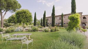 A garden with a fountain, tall trees, and a building with a brick facade. - Family Apartment  in the heart of Tuscany with swimming pool (Da Vinci) (cortona)