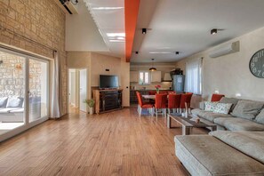 A spacious living area with a sofa, a dining table with chairs, a television, and a kitchen in the background.