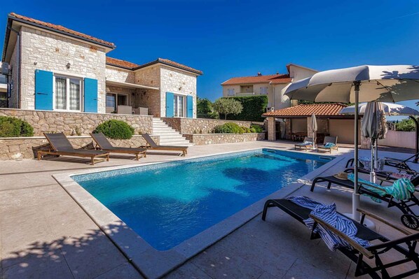 A poolside area with lounge chairs, a swimming pool, stone buildings with blue shutters, and a clear blue sky.
