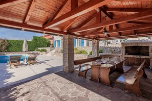 An outdoor patio with stone flooring, wooden furniture, a pool, and a stone fireplace.