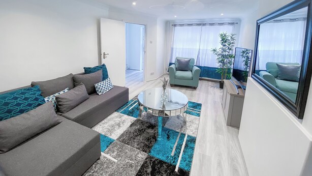 Ein modernes Wohnzimmer mit einem grauen Sofa, einem Glastisch und einem blauen Teppich mit Muster. In der Ecke steht ein grüner Sessel und ein Blumentopf.