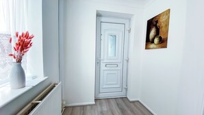 Ein Flur mit einer weißen Tür, einem Bild an der Wand und einem Fenster mit Blumen in einer Vase.