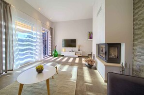 A modern living room with a flat-screen TV, fireplace, and large windows with blinds.