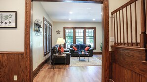 Una sala de estar con piso de madera, un sofá de cuero negro con cojines naranja y azules, una mesa de centro y una lámpara. Se observa una estantería con objetos decorativos y una ventana con vista a árboles.
