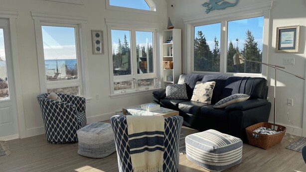 A spacious living room with a high ceiling, large windows offering a view of trees and the sky, a dark-colored sofa with patterned cushions, and a variety of seating options including ottomans and a chair.