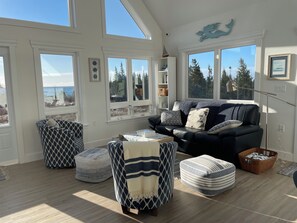 A spacious living room with a high ceiling, large windows offering a view of trees and the sky, a dark-colored sofa with patterned cushions, and a variety of seating options including ottomans and a chair. - Explore your own private beach and nature on 12 acres of ocean front property.  (Lake Charlotte)