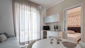 A modern hotel room with a bed, a TV, a dining table with placemats, and a large window with curtains. - Studio with community pool (Lido di Jesolo)
