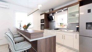A modern kitchen with a central island, bar stools, stainless steel appliances, and a window with flowers. - Villa with pool for 8 people (Malinska)