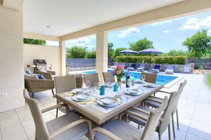 An outdoor dining area with a table set for four, surrounded by wicker chairs, a pool, and umbrellas in the background.