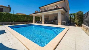 A rectangular swimming pool with a tiled deck, a covered patio area with outdoor furniture, and a two-story house with a balcony. - Villa with pool for 8 people (Malinska)