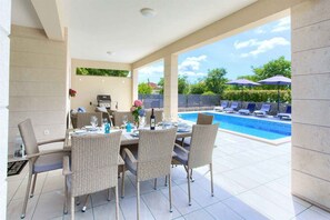 An outdoor dining area with wicker chairs and a table set for two, overlooking a swimming pool with lounge chairs and umbrellas.