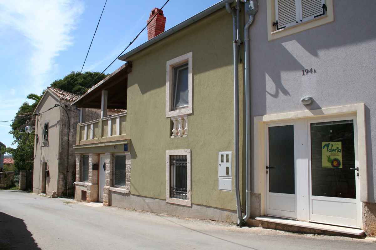 A two-story building with a green facade, a white door with a sign, and a small window with a metal grill.