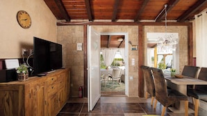 Un interior rústico con un mueble bajo para la televisión de madera, un comedor con sillas y vistas a un espacio exterior por una puerta abierta.