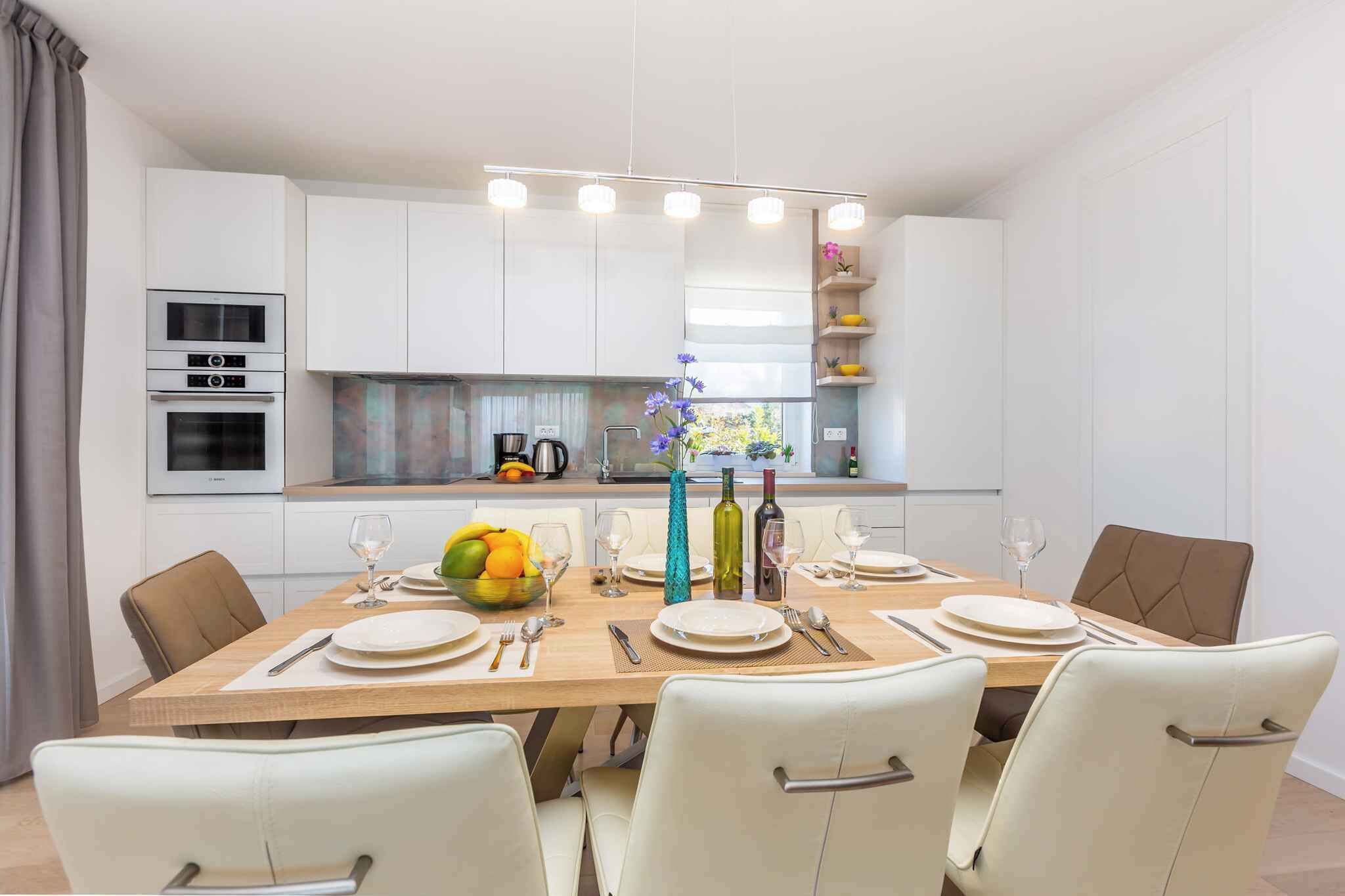 Un espace repas moderne avec une table en bois dressée pour six personnes, des chaises blanches et, au fond, une cuisine dotée de meubles et d’appareils électroménagers blancs.