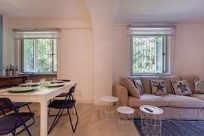 Una sala de estar moderna con comedor, ventanas amplias con rejas metálicas, un sofá beige con cojines estampados y una mesa de comedor blanca para cuatro personas.