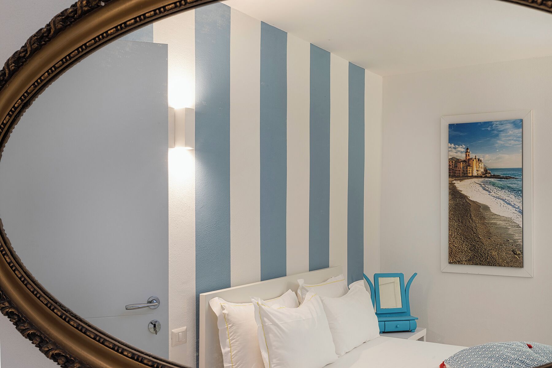 Une chambre d’hôtel avec un lit, un mur à rayures bleues et blanches, un tableau représentant une plage et un miroir doté d’un cadre orné.