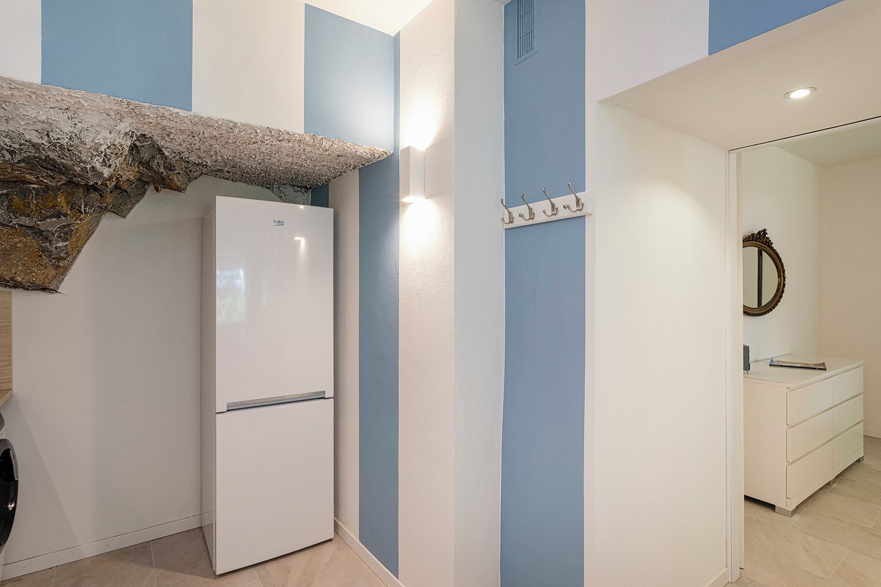A compact kitchen with a white refrigerator, a sink, and a mirror. The walls are painted in blue and white stripes.