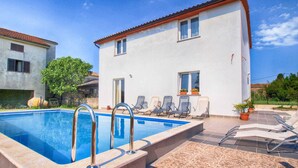 A swimming pool with lounge chairs, a white building with a red roof, and a tiled patio area.
