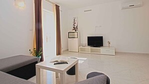 A modern living room with a sofa, a small table with a book and remote, a TV on a white stand, and a potted plant.