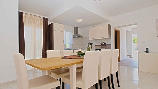 A modern dining area with a wooden table and white chairs, a kitchen in the background, and a view of the outdoors through large windows.