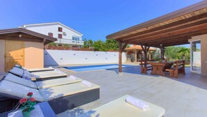 A poolside area with lounge chairs, a wooden pergola, and a view of a white building. - Holiday home with terrace for 2 people (Vantačići)