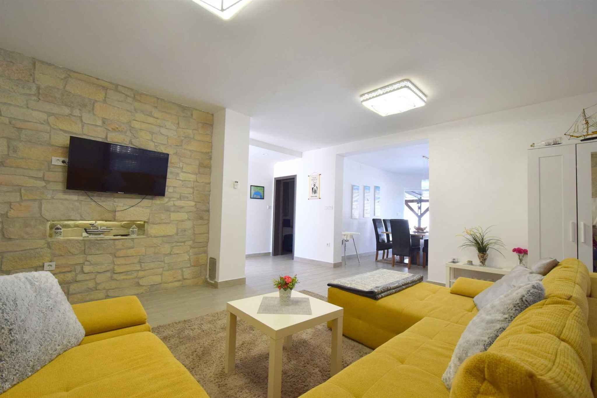 Uma sala de estar com parede de pedra, televisão de tela plana, sofás amarelos, uma mesa pequena com um vaso e uma área de jantar ao fundo.