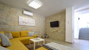 A living room with a yellow sofa, a white coffee table, a patterned rug, a stone wall, a flat-screen TV, and a staircase leading to an upper level. - Holiday home with terrace for 2 people (Vantačići)