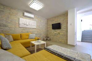 A living room with a yellow sofa, a white coffee table, a patterned rug, a stone wall, a flat-screen TV, and a staircase leading to an upper level.