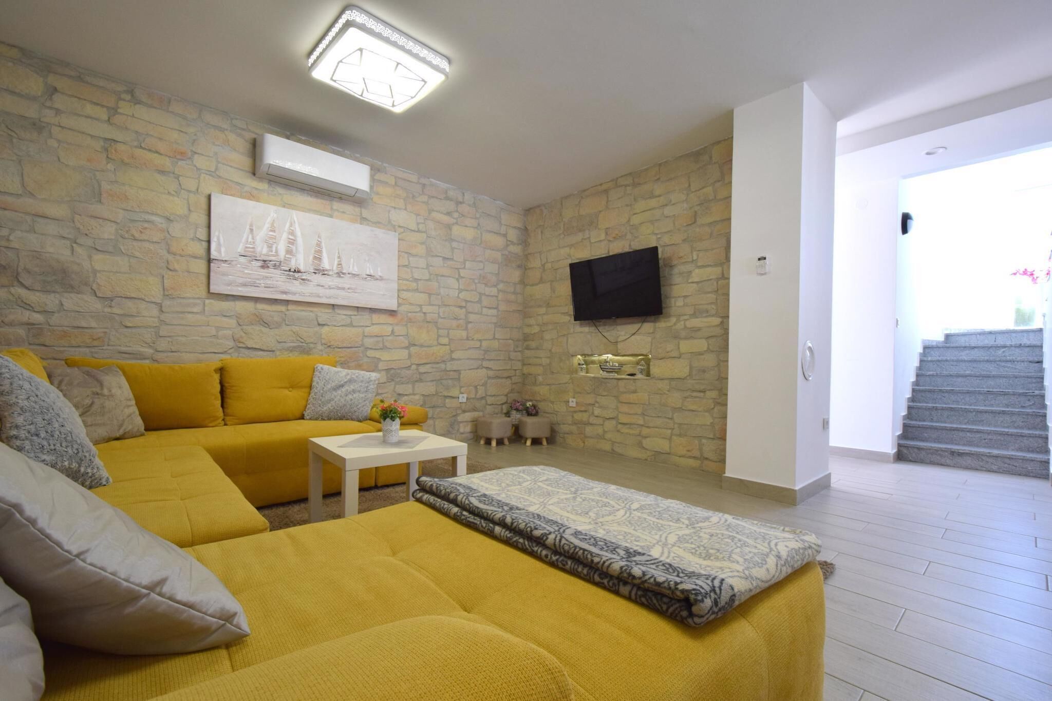 A living room with a yellow sofa, a white coffee table, and a patterned rug. There is a television mounted on the wall and a painting above it.