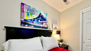 Un dormitorio con colcha roja, almohadas blancas, cabecera negra, una obra de arte en la pared, una lámpara de noche y una puerta.