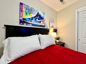 Un dormitorio con colcha roja, almohadas blancas, cabecera negra, una obra de arte en la pared, una lámpara de noche y una puerta.