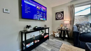 Un salón con un televisor de pantalla plana grande montado en la pared, una consola negra con objetos decorativos, una mesita con una lámpara y un sofá azul oscuro.