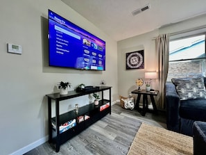 Un salón con un televisor de pantalla plana grande montado en la pared, una consola negra con objetos decorativos, una mesita con una lámpara y un sofá azul oscuro.