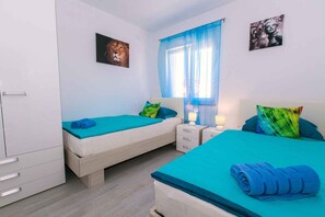 Two single beds with blue bedspreads and matching pillows, a large window, and two bedside tables with lamps.