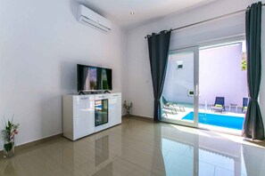 Un soggiorno moderno con una TV a schermo piatto, un mobile bianco e una porta in vetro che dà su una zona piscina.