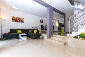 A modern living room with a black sectional sofa, a white coffee table, a painting on the wall, a dining area, and a staircase leading to an upper level. - Holiday home with pool and kilma system (Krk)