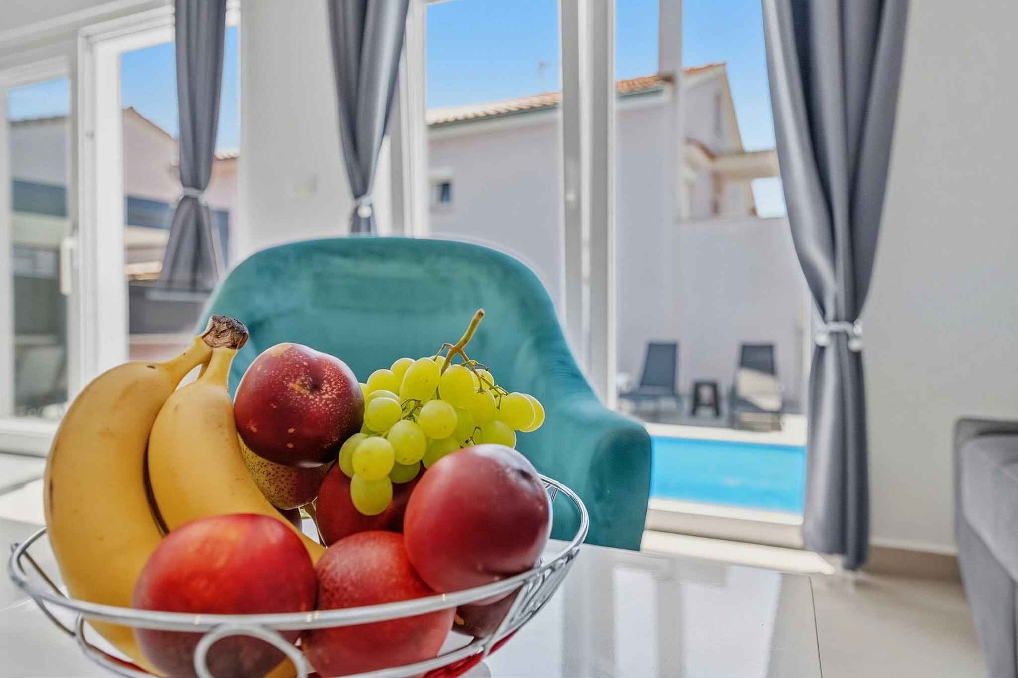 A room with a view of a pool, a teal chair, and a fruit bowl on a table.