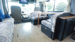 Interior compacto de una autocaravana con sofá, mesa de comedor, televisor montado en la pared y ventana con vistas al exterior.