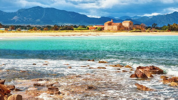 A coastal scene with a beach, clear turquoise waters, rocky shoreline, and a building with a red roof in the background. - Studio with swimming pool, near the sea (Pula Sardinien)