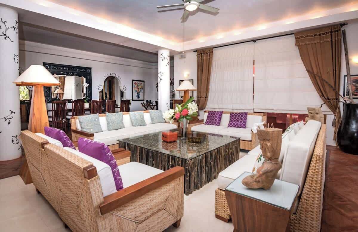 A spacious living room with a large sofa, a coffee table, and wicker furniture. There are lamps, a statue, and a large window with curtains.