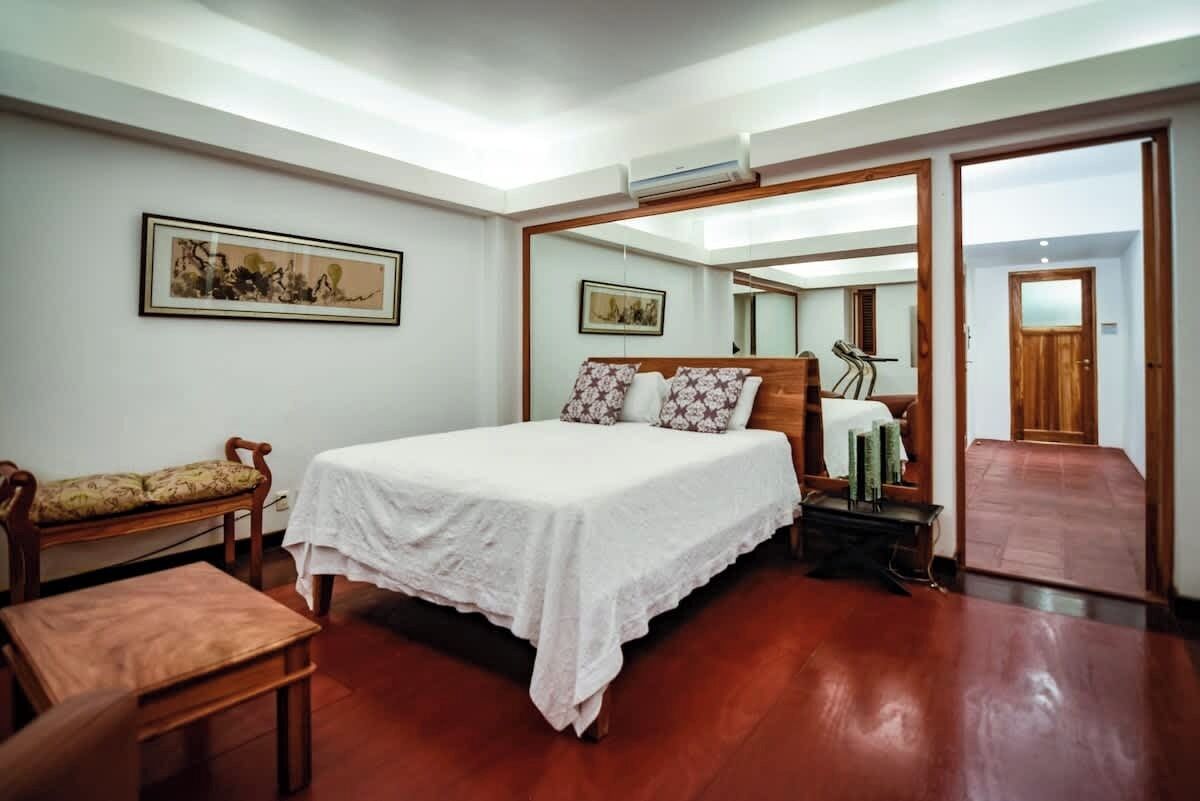 A bedroom with a large bed, a wooden bench, a small table, and a mirror.