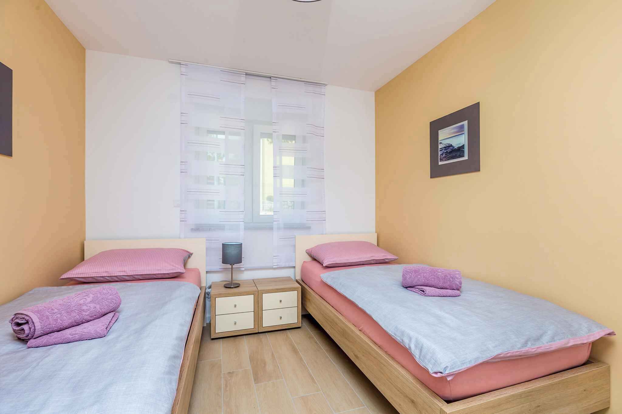 Une petite chambre bien éclairée, avec deux lits simples, une table de chevet et une fenêtre ornée de rideaux légers.