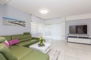 A modern living room with a green sofa, a coffee table with a plant, a flat-screen TV, and a large window with blinds.