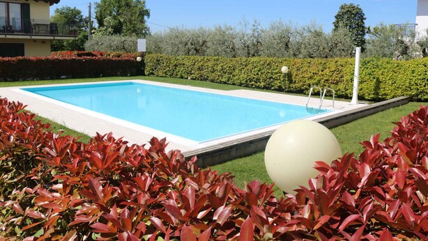Una piscina rectangular con un suelo blanco, rodeada de setos y césped cuidados.