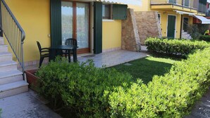Un edificio amarillo con una escalera, una puerta con contraventanas verdes y un pequeño patio con una silla y una mesa.